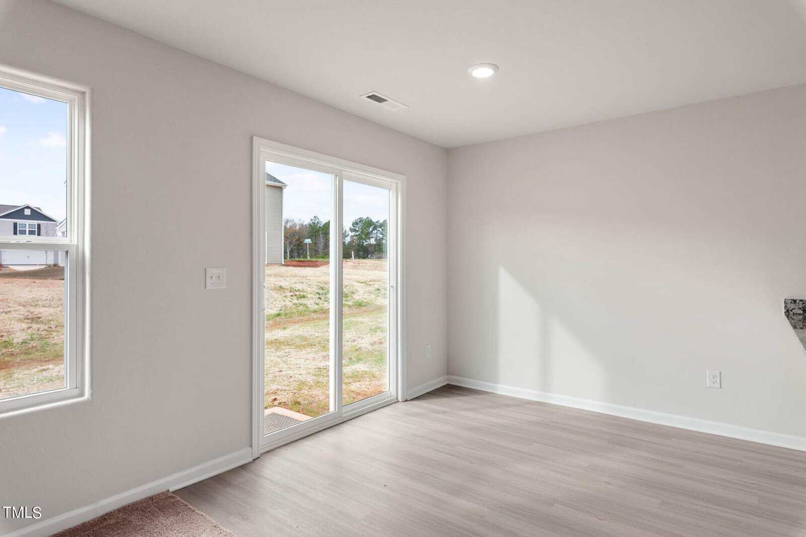 Empty room with light gray walls and wood flooring, featuring a large glass sliding door leading to a grassy outdoor area. Ceiling light is on, and there are no furnishings in the room.