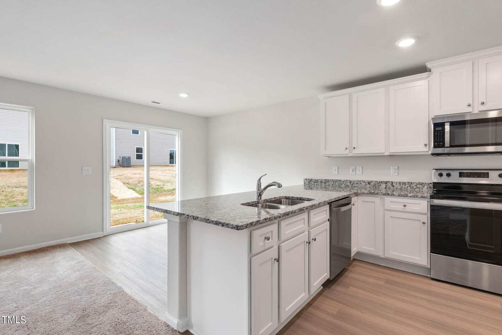 Modern kitchen with white cabinets, stainless steel appliances, and a granite countertop island with a sink. Hardwood floors are visible, and a sliding glass door opens to a backyard, providing natural light.