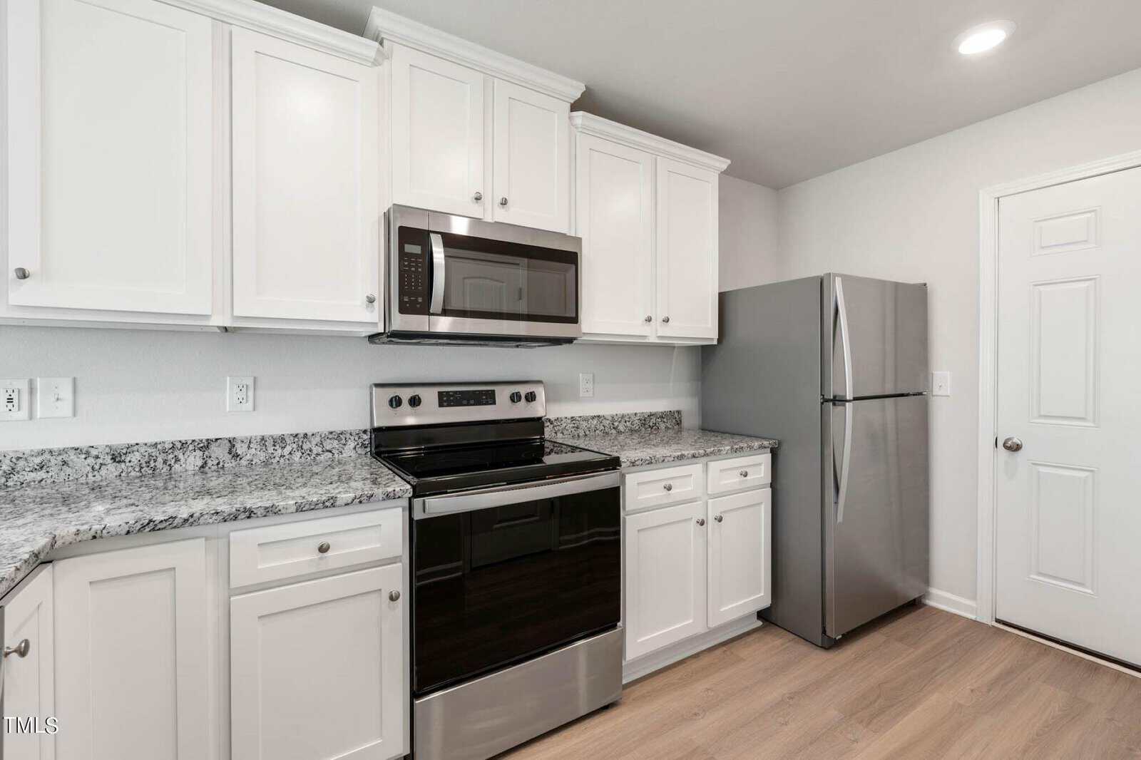 A modern kitchen with white cabinets, a granite countertop, a stainless steel stove with an overhead microwave, and a refrigerator. The floor is light wood, and there's a closed white door on the right.