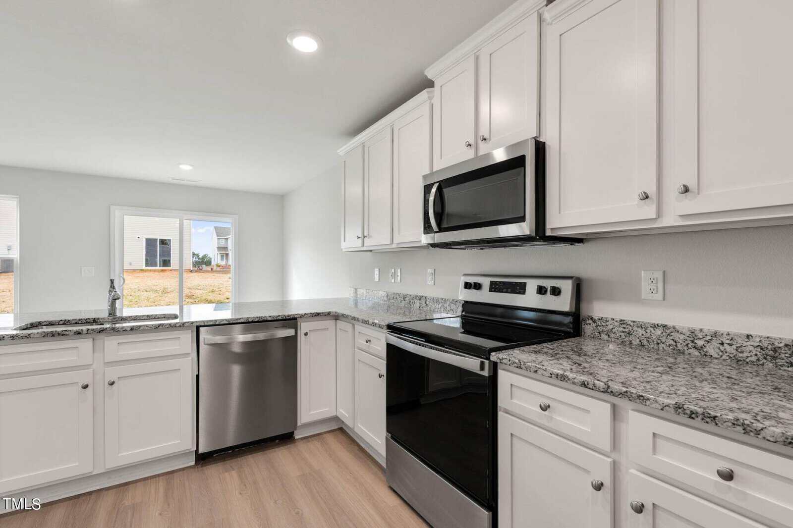 Modern kitchen with white cabinets and granite countertops. It features stainless steel appliances including a dishwasher, oven, and microwave. Light wood flooring and recessed lighting create a bright and open atmosphere. Large window offers natural light.