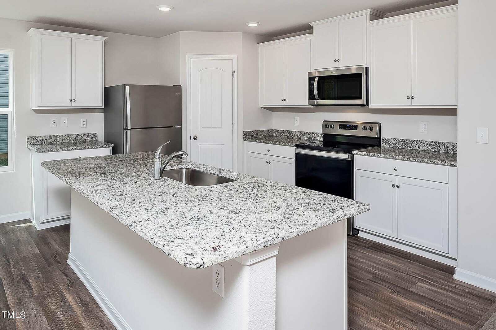 A modern kitchen with granite countertops, white cabinets, and stainless steel appliances, including a refrigerator and a stove. There's a large central island with a sink and faucet, and the floor is wood.