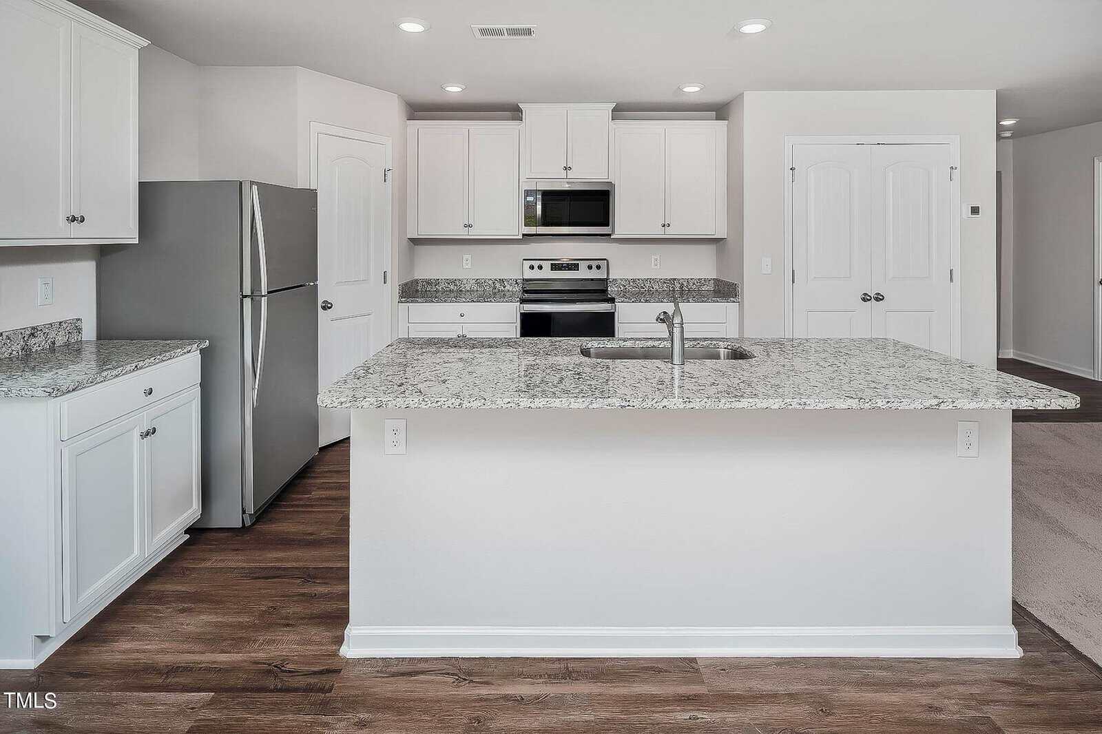 Modern kitchen with white cabinets, stainless steel appliances, and granite countertops. A large island with a sink is centered, and the floor features wood-style vinyl. Background includes a refrigerator, stove, and microwave.