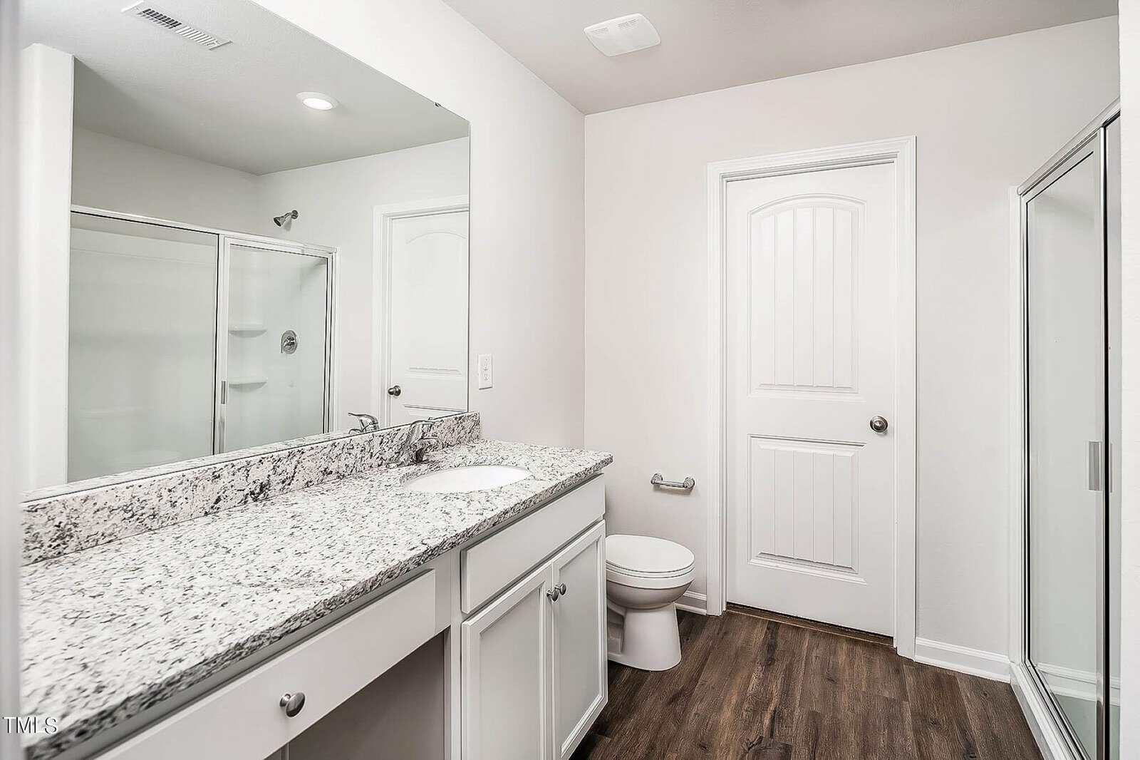 A modern bathroom features a granite countertop with a sink, white cabinets, a toilet, and a shower enclosure. The room has light-colored walls, and the floor is made of dark wood-style laminate. A white door leads to an adjacent space.