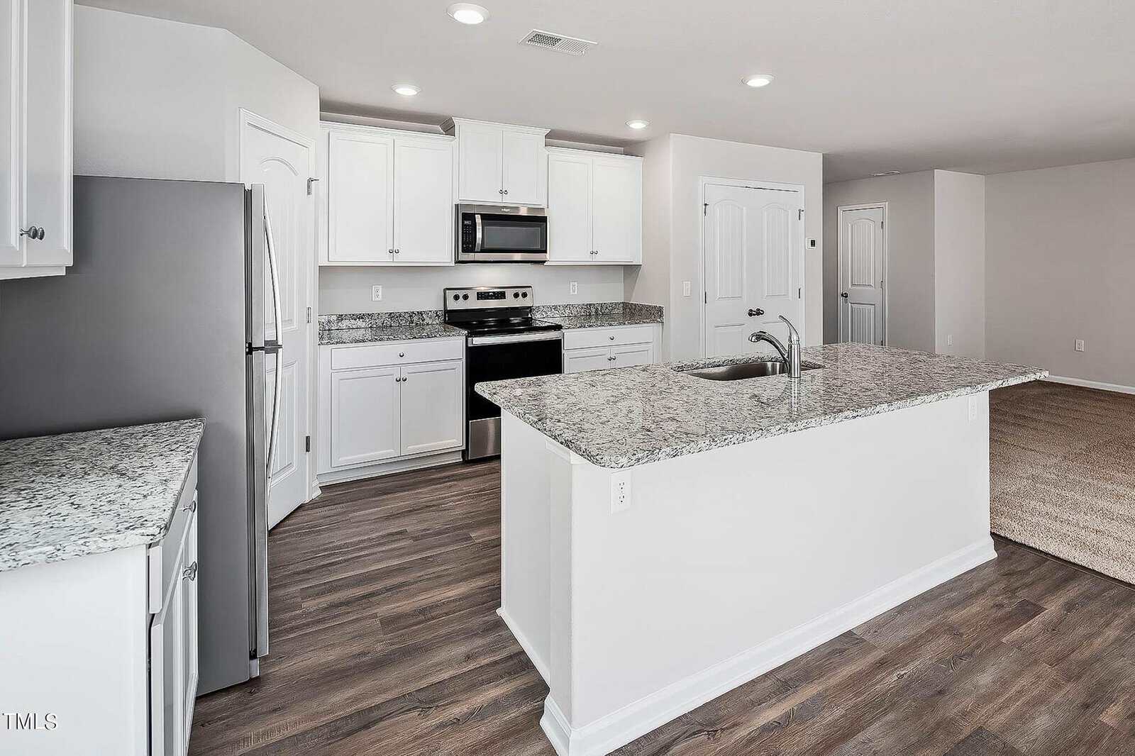 Modern kitchen with white cabinets, granite countertops, and stainless steel appliances, including a stove and microwave. A large island with a sink is centered on wood laminate flooring, with white walls and doors completing the look.
