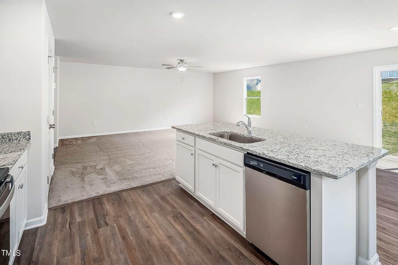A modern kitchen with white cabinets, a granite countertop island with a built-in sink and dishwasher. The space opens to a carpeted living area with a ceiling fan and a window, allowing natural light to enter.