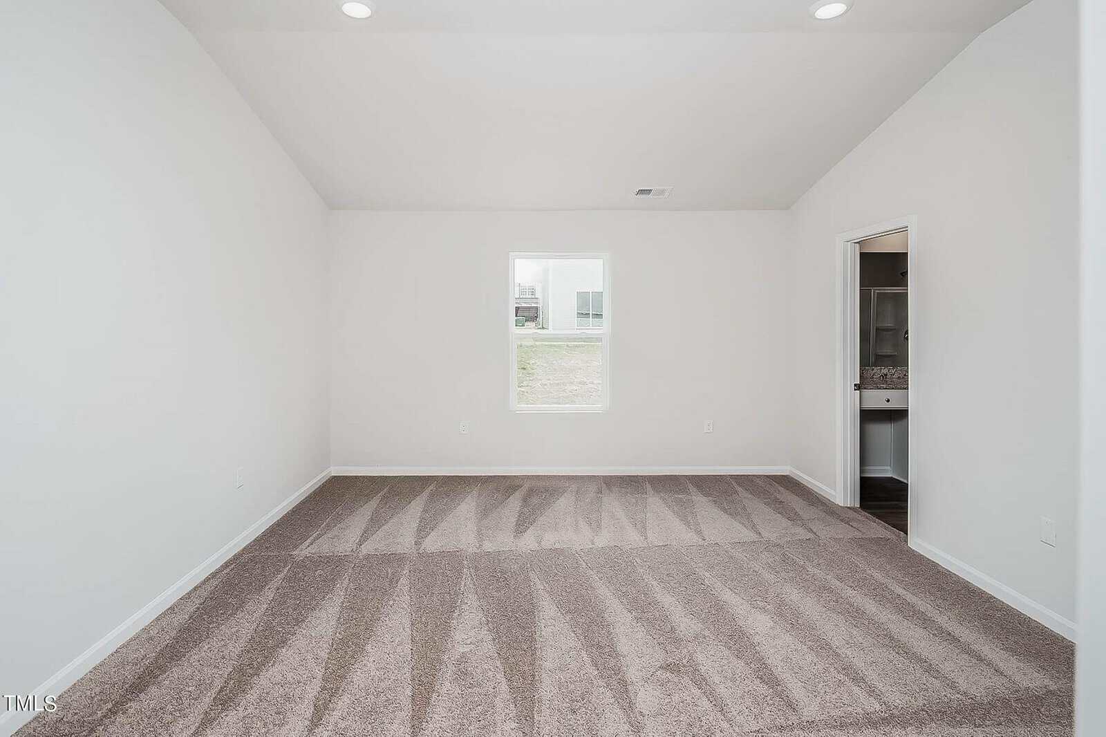 A bright, empty room with a high ceiling and neutral-colored carpet. The room features a central window with a view outside, and an open doorway leading to a bathroom with visible fixtures. Walls are painted white.