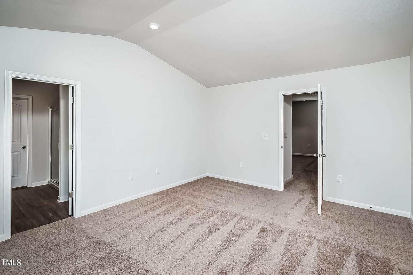 An empty, well-lit room with white walls and beige carpet. Two open doors reveal additional rooms with similar carpets. The ceiling has a recessed light, and the room appears spacious and clean.