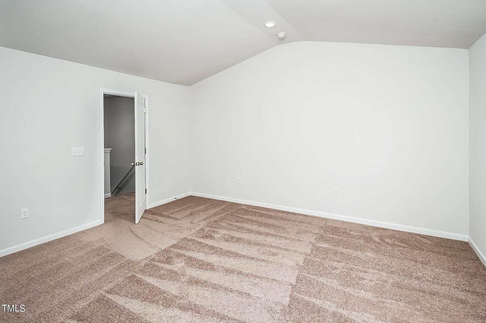 A spacious, empty room with light beige carpet and plain white walls. The room has an open door leading to a hallway with a visible staircase railing. Ceiling is slightly vaulted. There's a single ceiling light fixture.