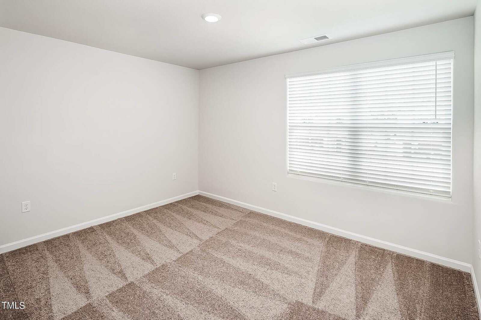 An empty room with beige carpet, white walls, and a window with blinds. Natural light fills the space through the window, creating a bright and clean atmosphere.