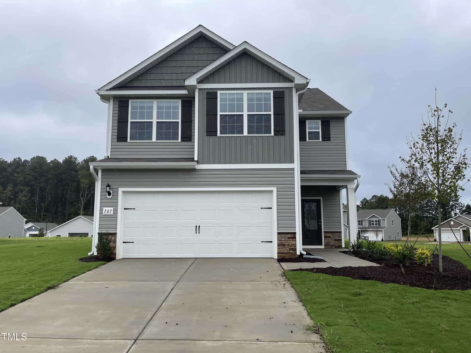 The charming two-story house at 802 Rhino Bend, Oxford, boasts gray siding and white trim, complete with a double garage door. The front yard is beautifully landscaped with lush green grass and a young tree, all set against an overcast sky backdrop.