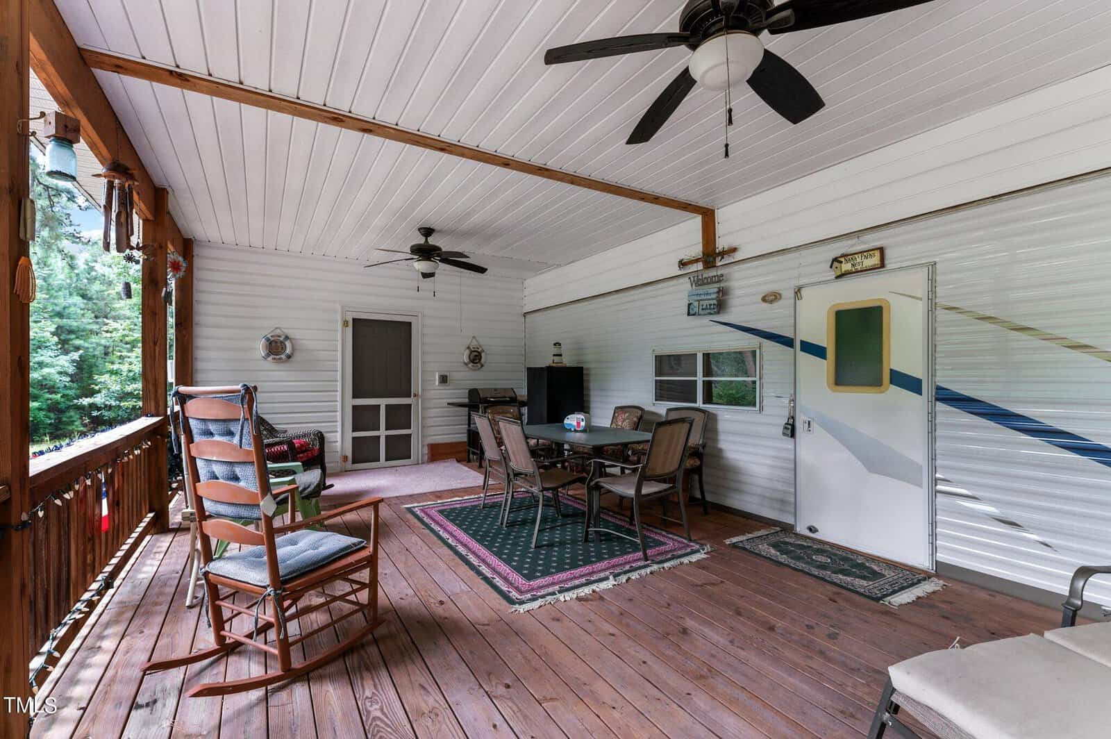 A covered porch with a wooden floor, ceiling fans, rocking chairs, a table with four chairs on rugs, and a door leading into a camper trailer. Trees are visible through the porch railing.