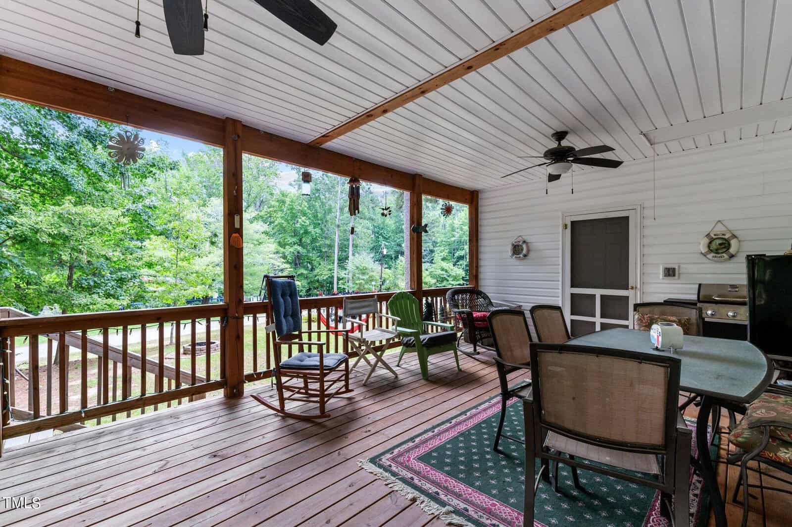 Covered wooden porch with ceiling fans, various chairs, a grill, table with chairs, and decorative items. The porch overlooks a green, wooded backyard. Daylight streams in through the open sides.