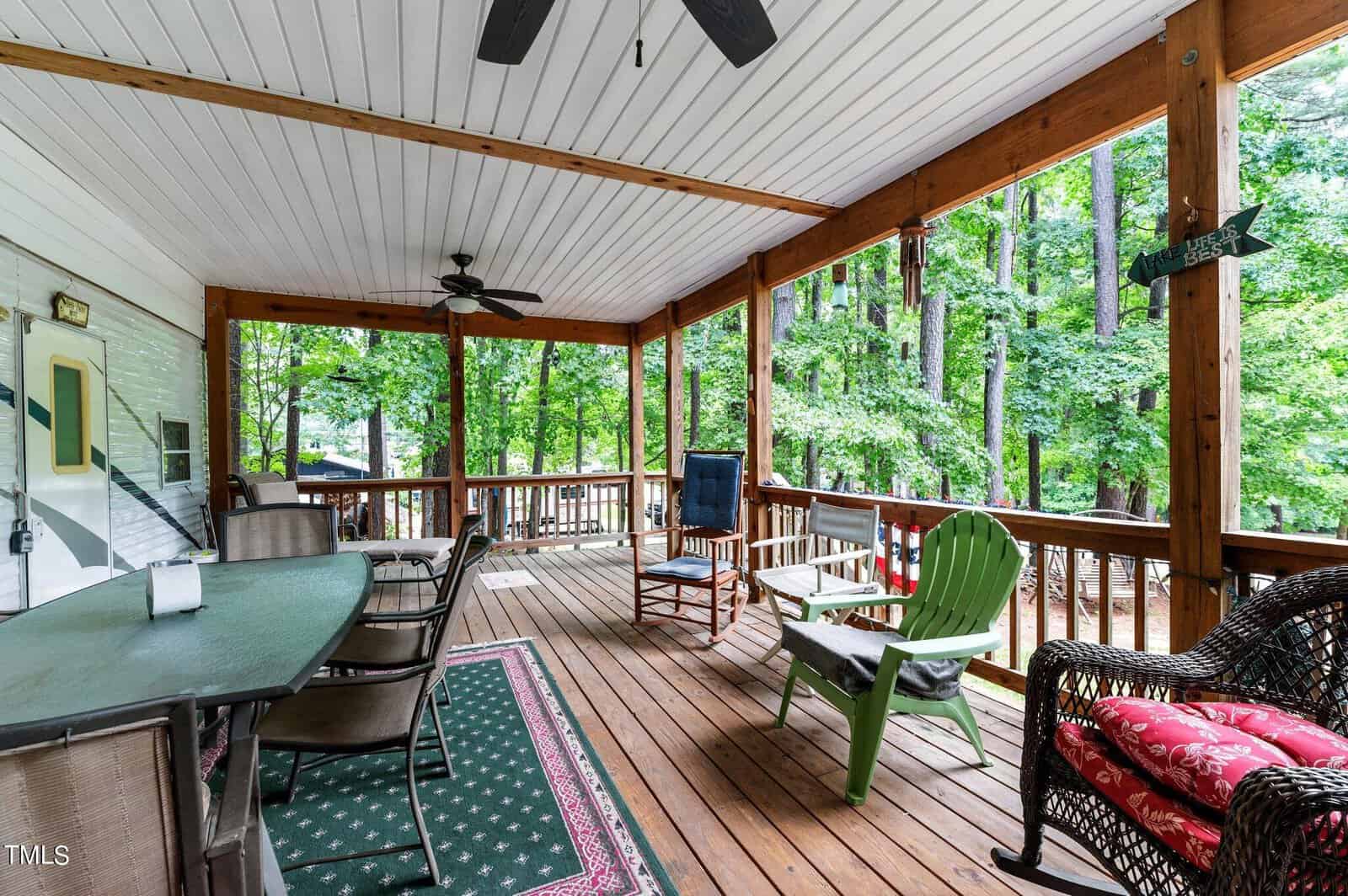 Covered wooden porch with ceiling fans, patio furniture including a dining table, chairs, and armchairs, surrounded by trees and greenery, creating a cozy outdoor living space.
