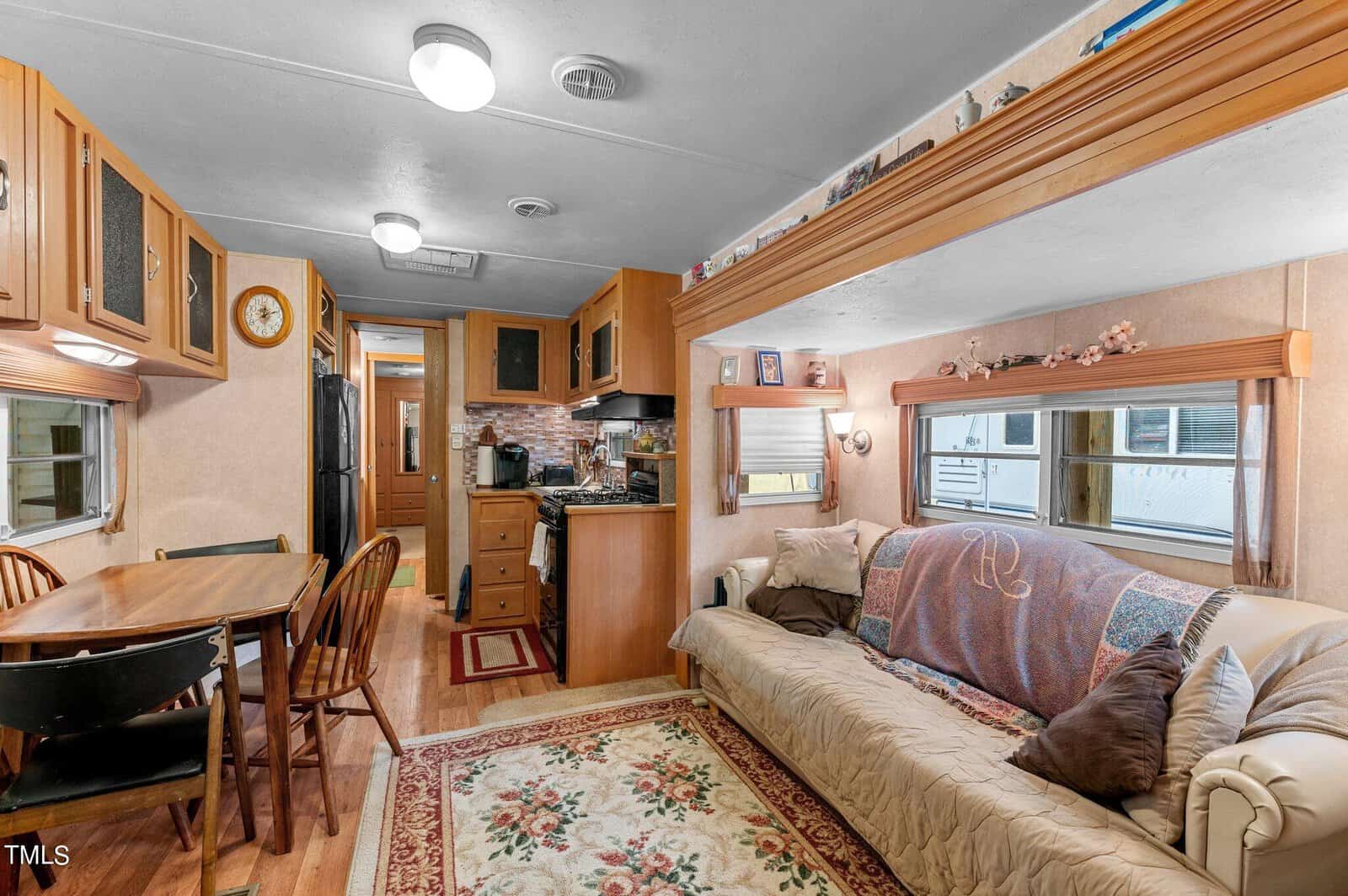 Interior of a cozy RV with a dining table and chairs on the left, a kitchen with wood cabinets and appliances in the center, and a beige sofa with pillows and a blanket on the right, all set on a rug with decorative accents.