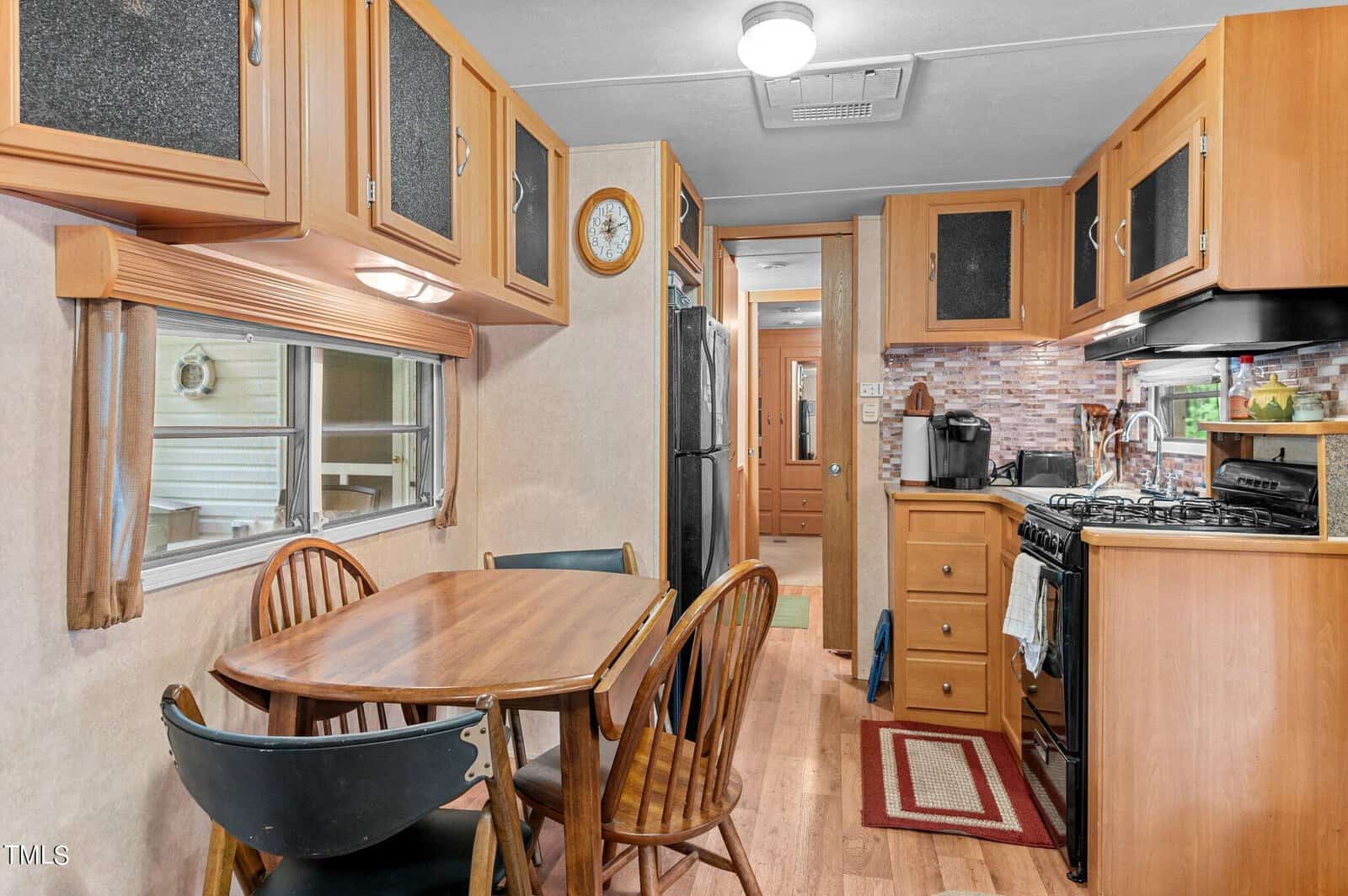 A cozy RV kitchen and dining area with a wooden table and chairs, wood cabinets, a stove, microwave, fridge, and a window with a curtain. The kitchen has a tile backsplash and a coffee maker on the counter.