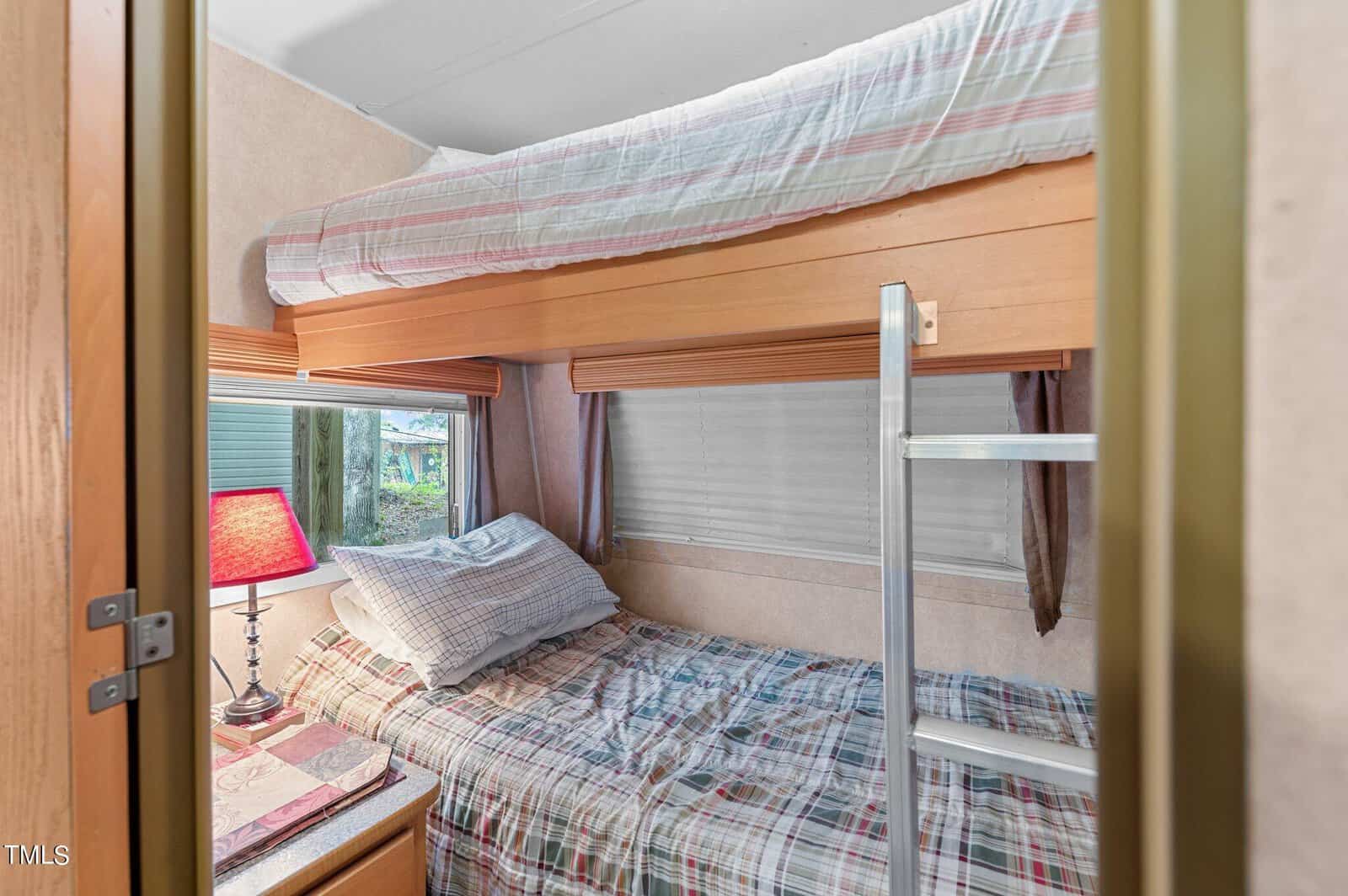 Small room with wooden bunk beds, plaid bedding, a silver ladder, and a window. A bedside table with a pink lamp and books is next to the lower bed.