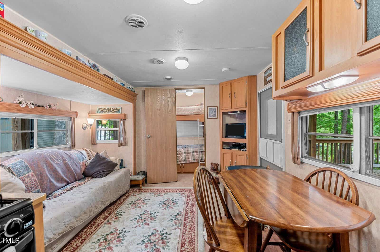 A cozy RV interior featuring a wooden dining table with chairs, a couch with a blanket, a patterned rug, built-in cabinets, large windows, and a view into a bedroom with a small TV and bunk beds in the background.