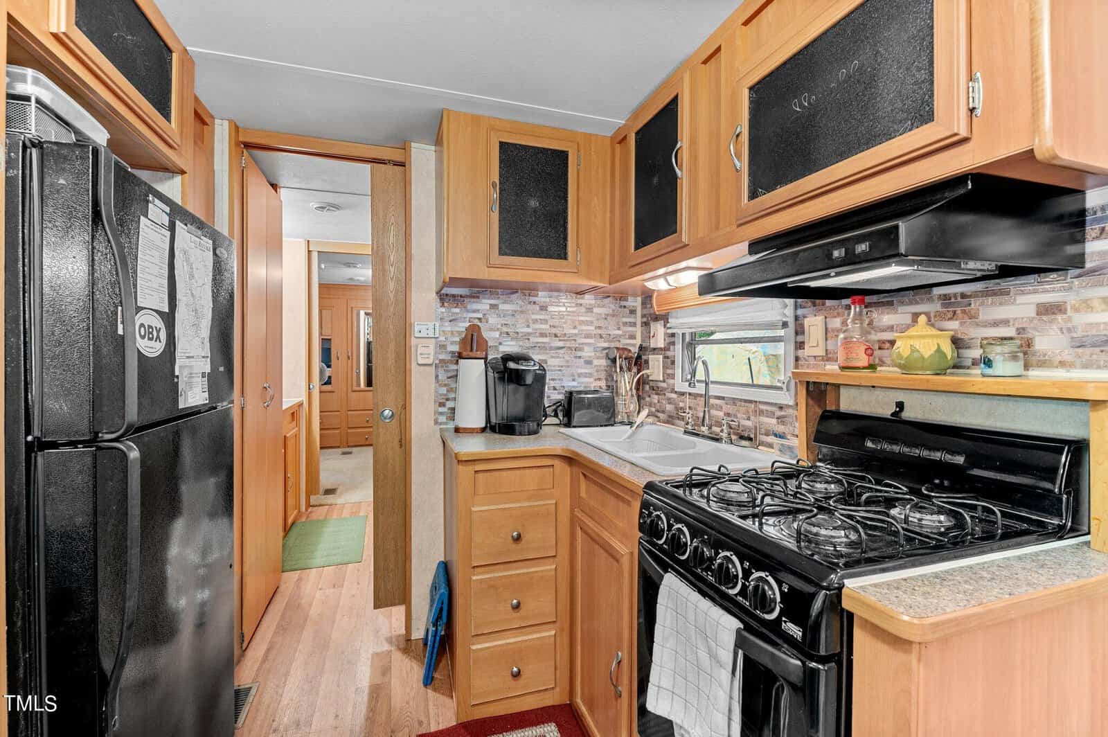 A compact RV kitchen with wooden cabinets, a black refrigerator, gas stove, oven, sink, coffee maker, and a tile backsplash. A hallway leads to another room, and natural light enters through a small window.