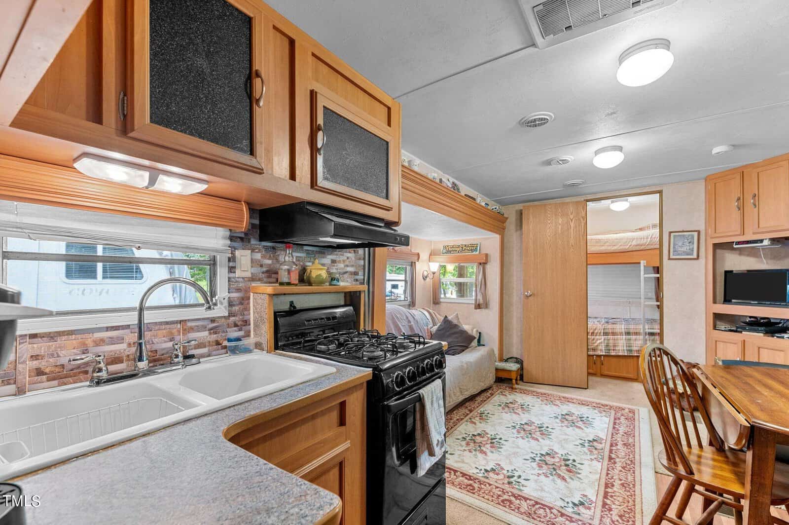 Interior of an RV with a kitchen, stove, sink, sofa, dining table with chairs, TV, bunk beds, and a patterned rug. Wood cabinets and windows provide a cozy, home-like feel.