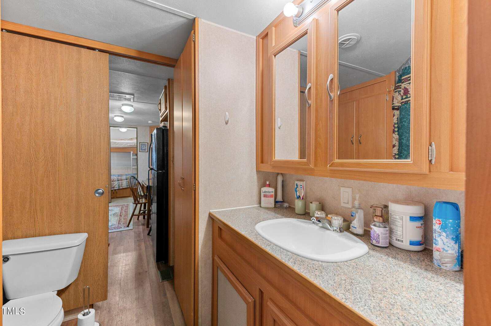 Bathroom with a wooden cabinet vanity, sink, and toiletries on the counter. A toilet is to the left. A hallway leads to another room with visible furniture and a bed in the background.