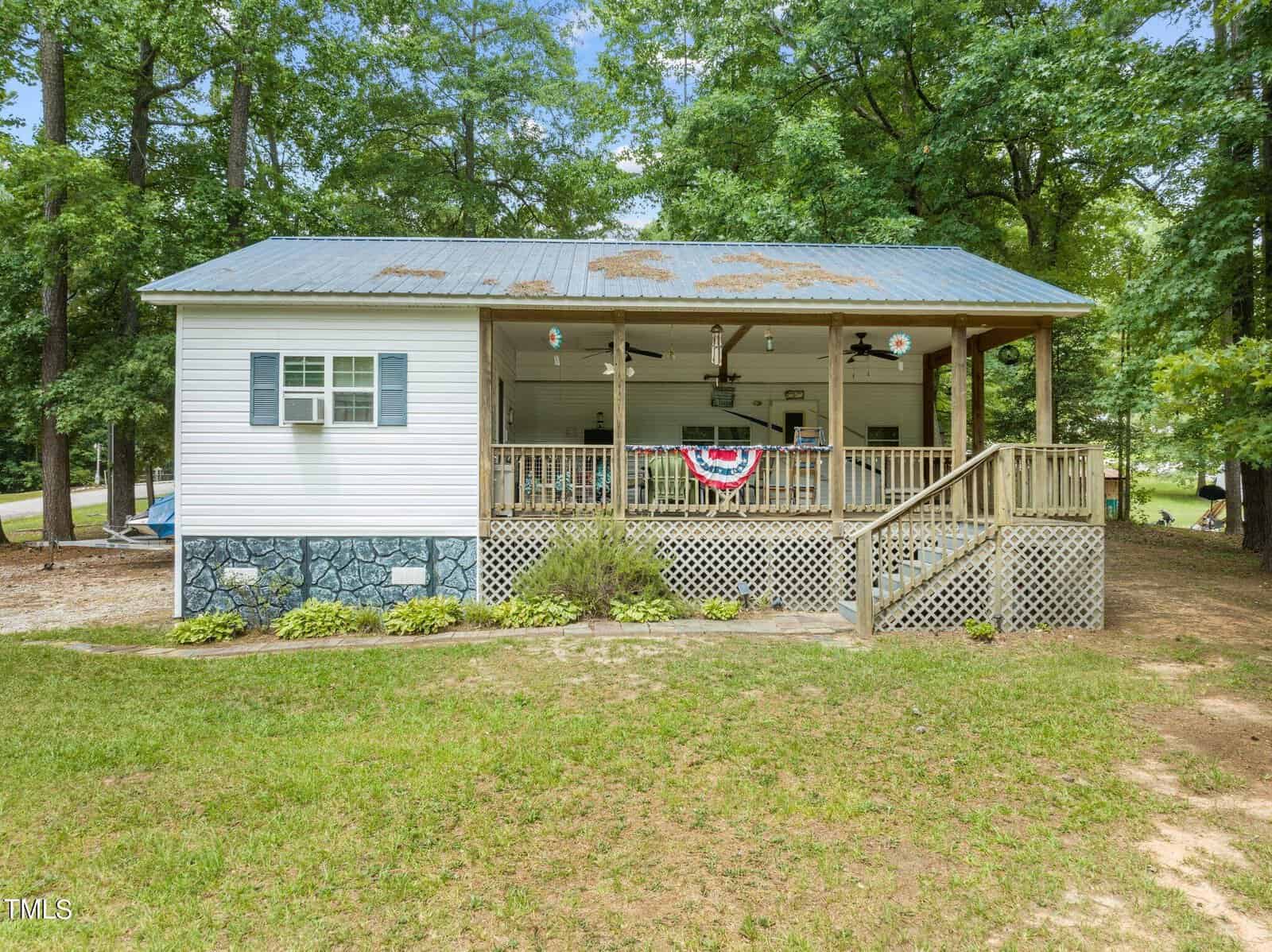 A small white house with a metal roof, front porch, and wooden steps, surrounded by trees and grass. The porch is decorated with a red, white, and blue banner; the house has an air conditioner in the window.