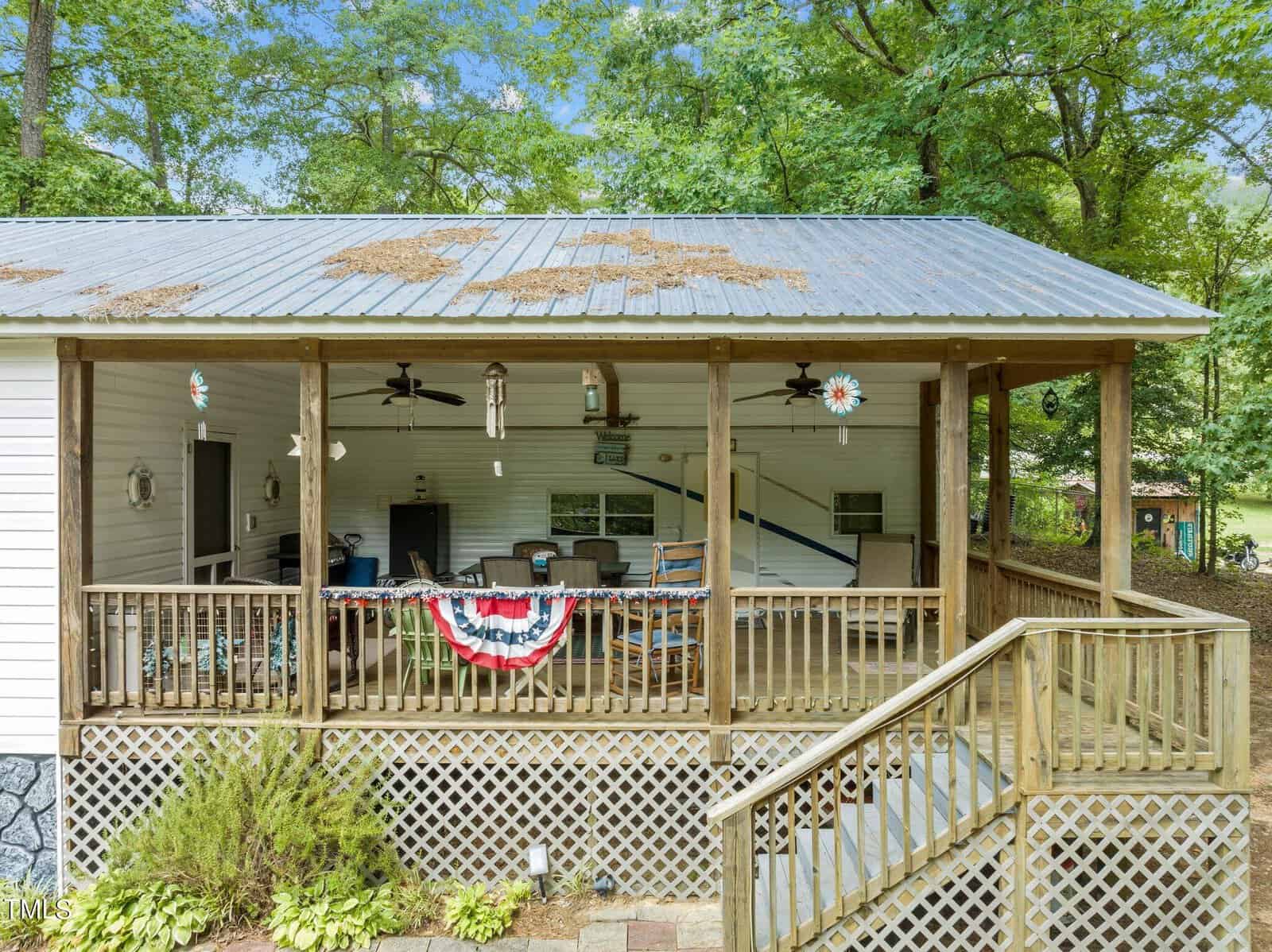 A covered wooden porch with a metal roof, ceiling fans, wind chimes, and patriotic bunting. Outdoor furniture and a hammock are visible, surrounded by trees and greenery. Steps lead up to the porch.