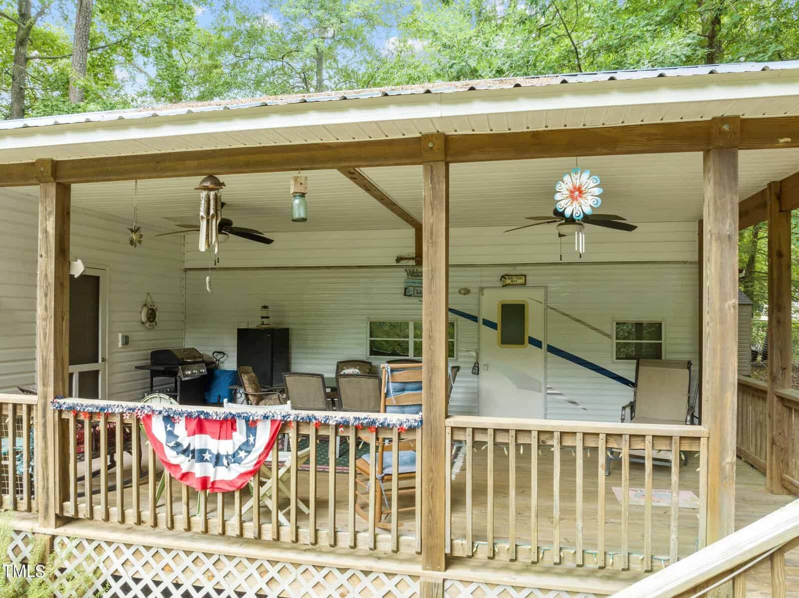 Covered wooden porch with ceiling fans, chairs, a grill, and a TV. Patriotic red, white, and blue bunting hangs on the railing. Trees and greenery surround the porch, which is attached to a white-sided building.