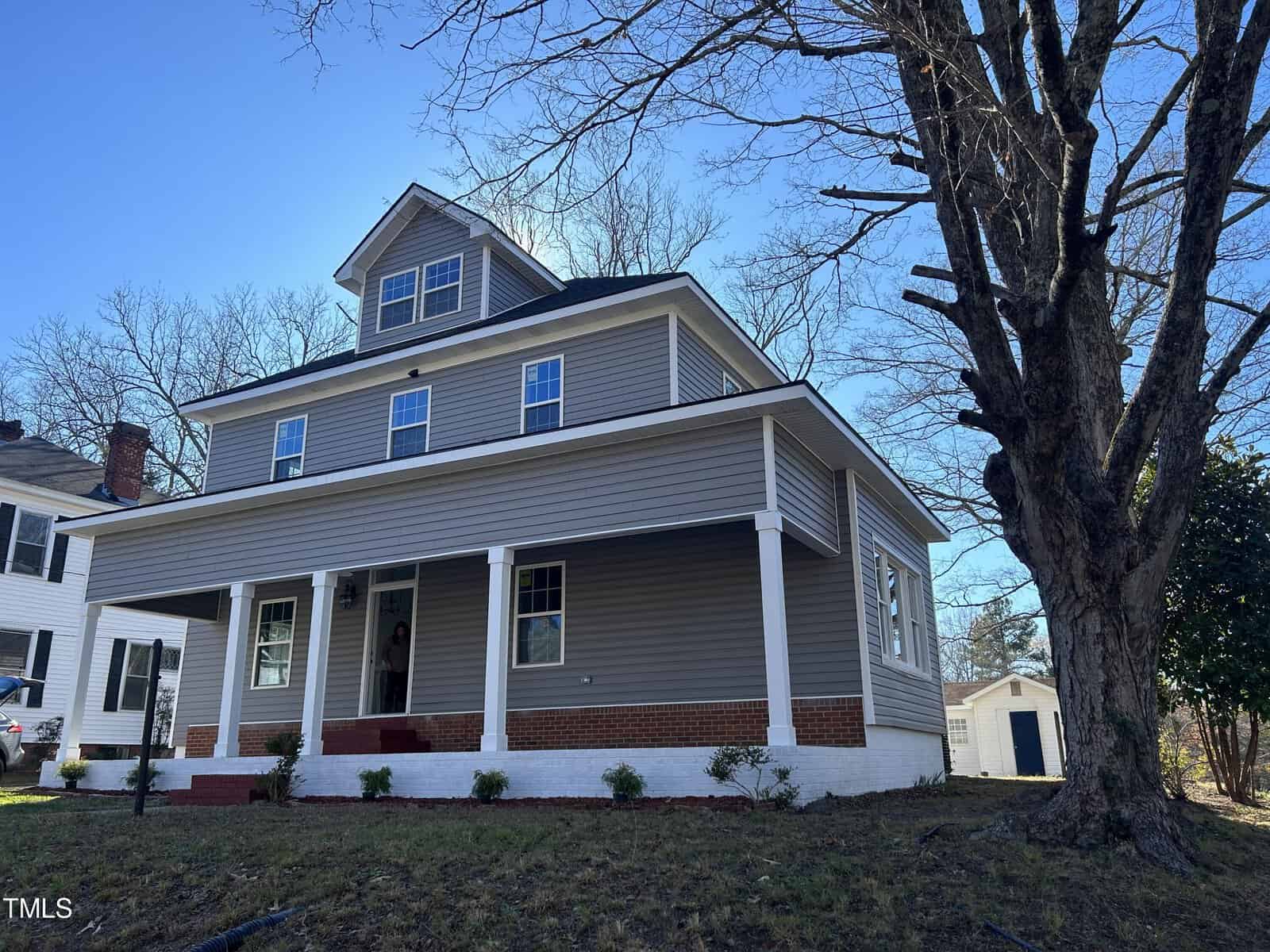 A two-story house at 243 Burwell Avenue in Henderson, NC, features gray siding and white trim. A spacious porch wraps around the front, where a person stands at the entrance. A large bare tree is in the foreground. The sky is clear and blue, with another house and a detached garage visible in the background.