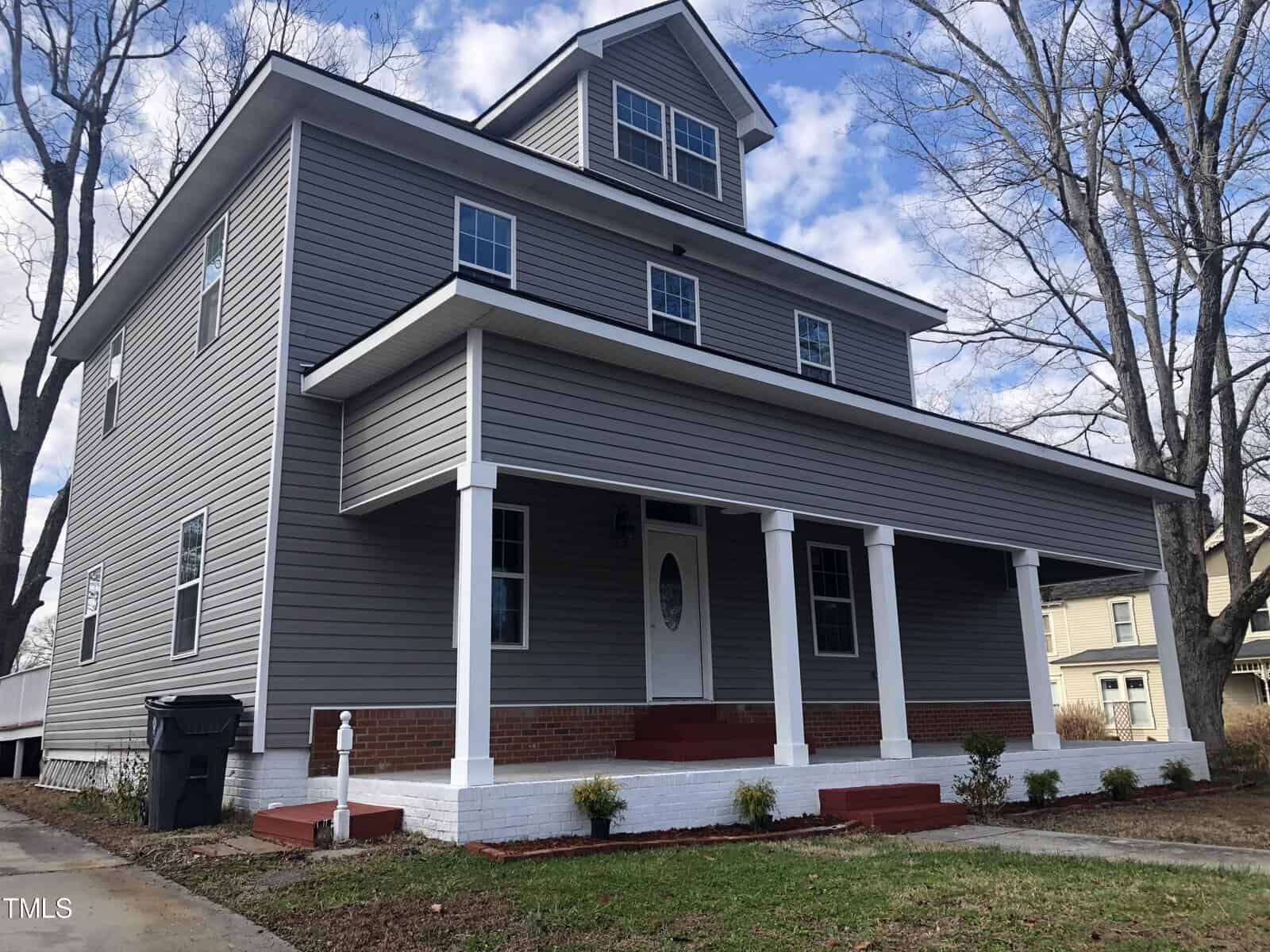 A two-story house with gray siding, white trim, and a covered front porch is seen on a sunny day. The house features multiple windows and an attic with additional windows. In the foreground, there is a garbage bin on the left side of the property and some small bushes.