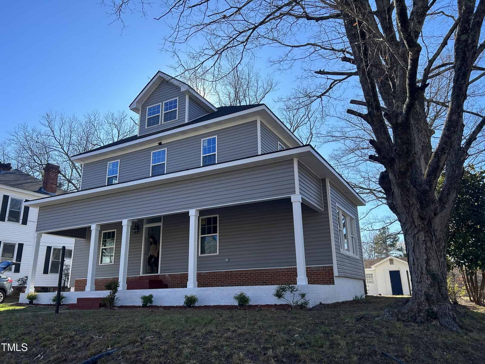 A two-story grey house with white trim and a front porch supported by white columns. The yard features grass and a large tree on the right. A person is visible on the porch, and the sky is clear with visible tree branches. Another house is partially visible to the left.