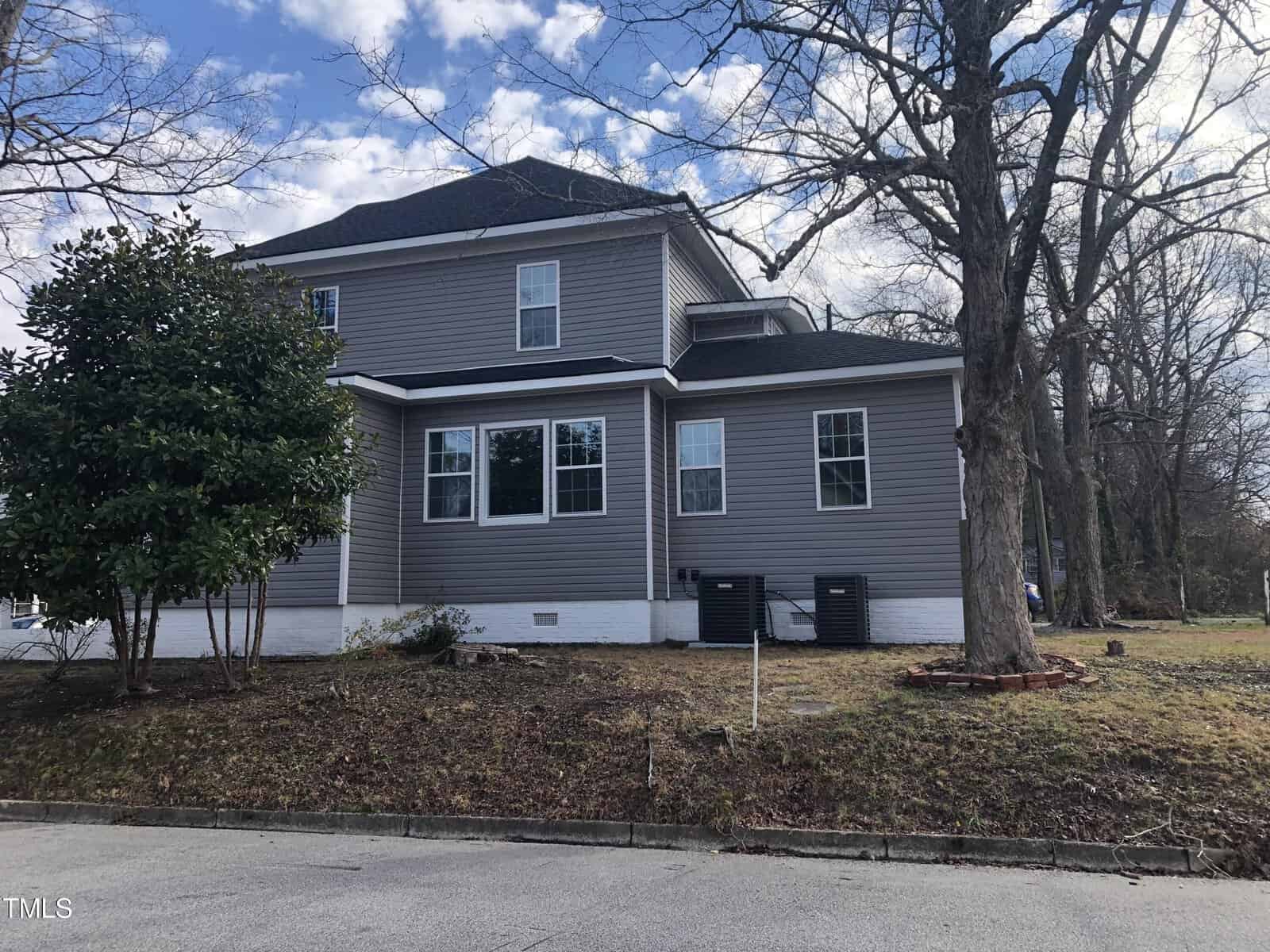 A two-story gray house with white trim, surrounded by leafless trees and sparse shrubbery. The lawn is brown, indicating it is likely winter. Two outdoor air conditioning units are visible on the side of the house. The sky is partly cloudy.