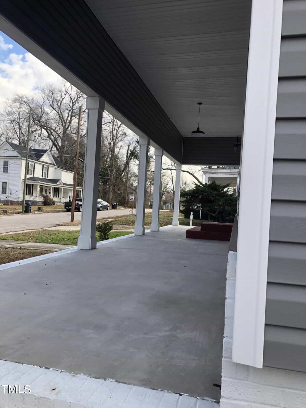 A wide, covered porch with white pillars and a gray, concrete floor. Gray siding walls with white trim are visible. The porch overlooks a street with some trees and neighboring houses in the background. A red brick step leads up to the porch.
