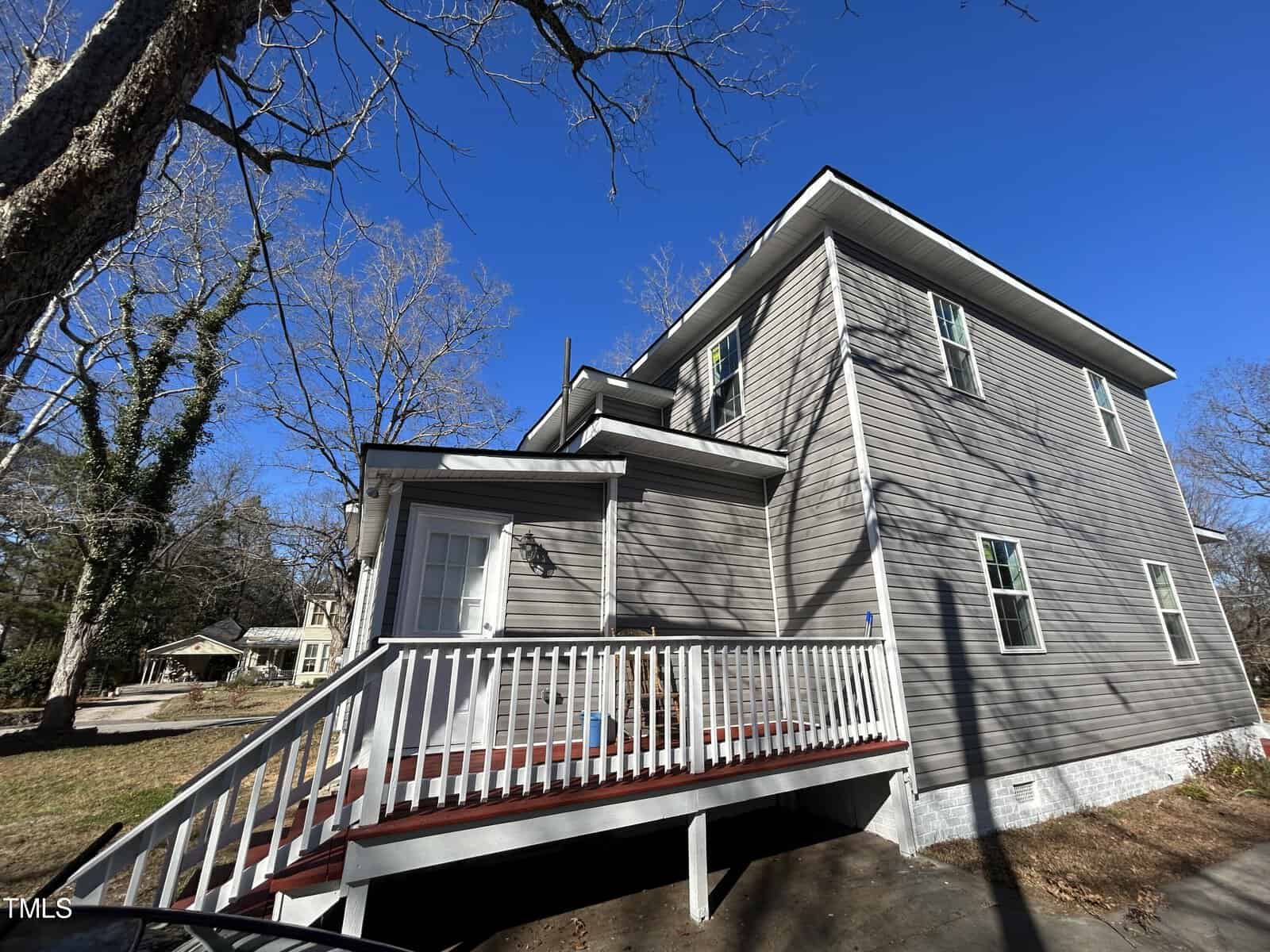 Two-story gray house with a small front porch and white railing in a suburban neighborhood. The house is surrounded by leafless trees under a clear blue sky. A driveway is visible, and other houses are seen in the background.