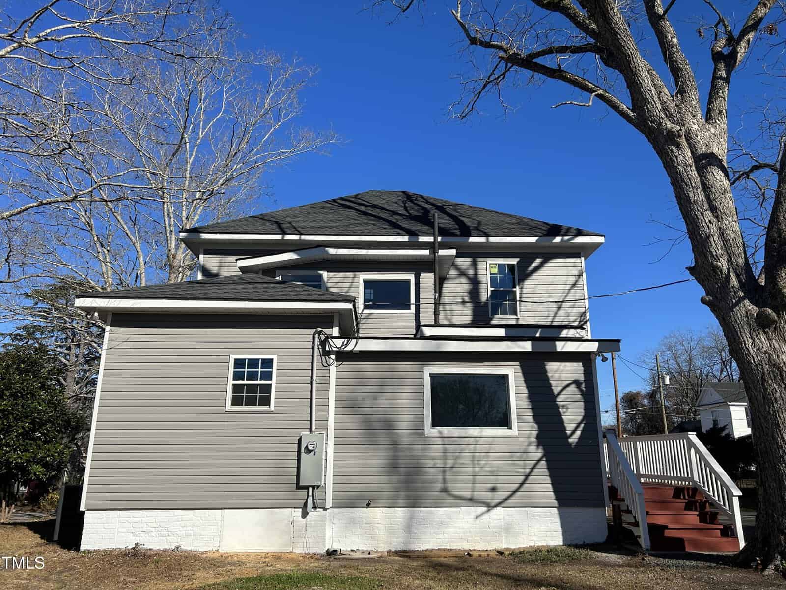 A two-story gray house with white trim on a sunny day. The house features black shingled roofing, an outdoor staircase on the right side, two prominent leafless trees, and a small patch of grass in the foreground. Clear blue sky in the background.