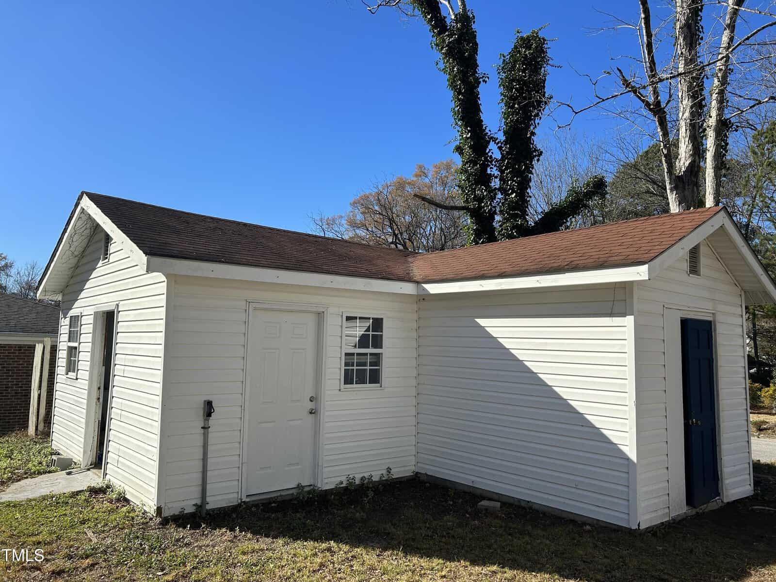 A small white shed in a yard with two doors (one white and one blue), a window, and a brown shingled roof. The shed has siding, and there's a large tree with ivy behind it. The sky is clear and blue.