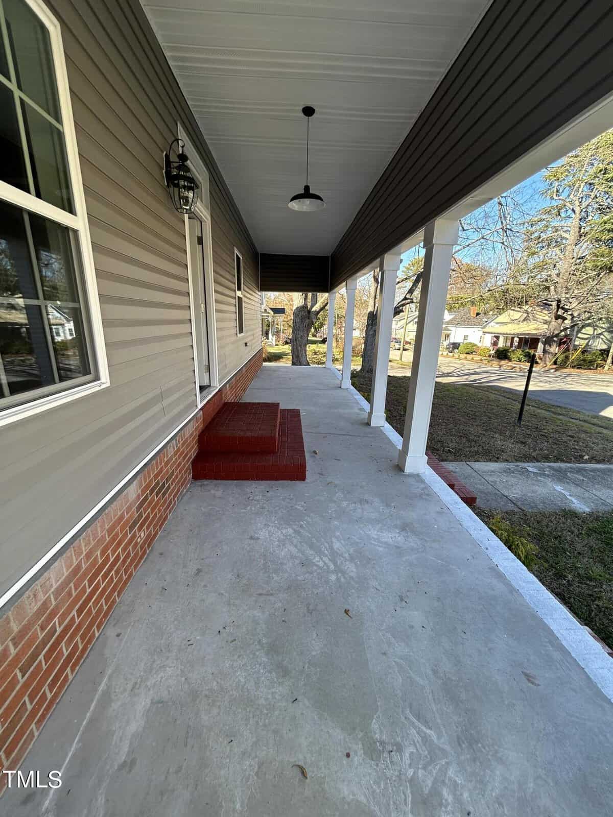 A spacious front porch with a cement floor, brick steps leading to the entrance, and white pillars supporting the roof. The house exterior is a combination of tan siding and red brick at the base. Trees and a residential street are visible in the background.