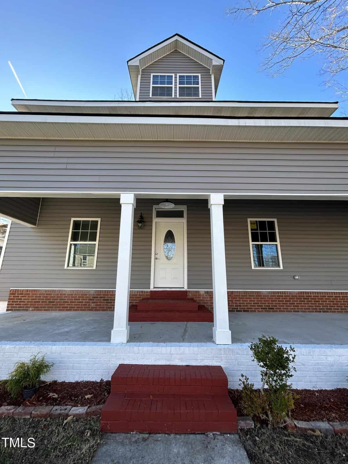 A front view of a two-story house with gray siding and white trim. The entrance features a white door with an oval window, red steps, and a brick foundation. Two white columns support the roof over a small porch. The sky is clear and blue.