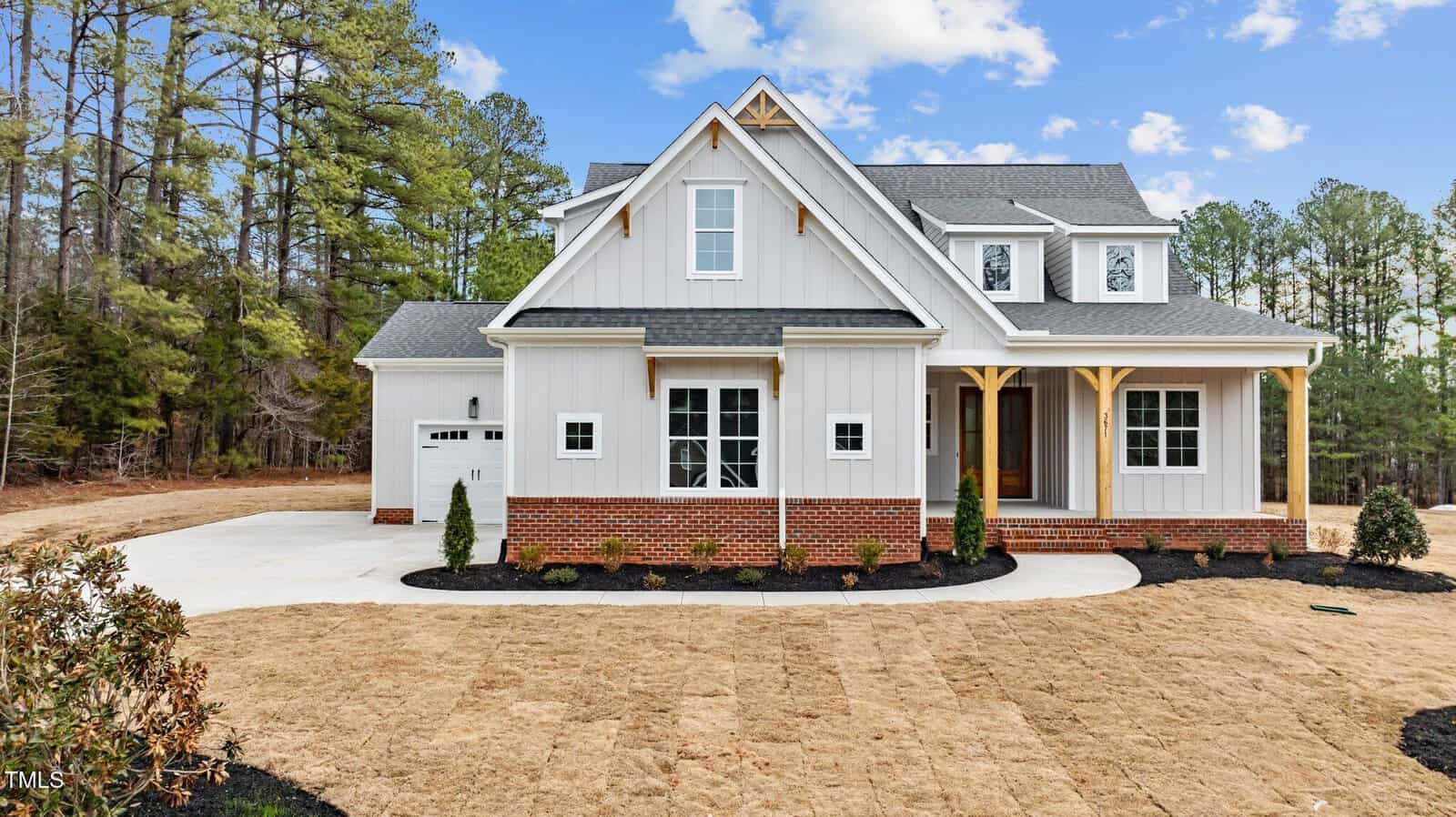 A modern gray house with a steep gable roof, white trim, and a front porch. Brick accents are at the base. The driveway is on the left, and the yard is covered with patchy brown grass. Surrounding the house are tall trees under a blue sky with clouds.