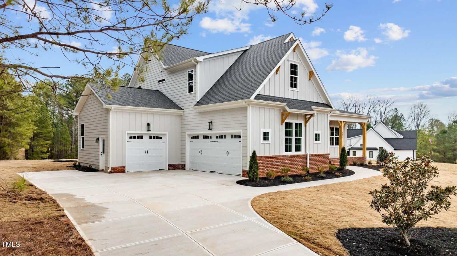 A modern two-story house with white siding and brick accents, featuring two garage doors on the left. The front yard includes a curved driveway and a small landscaped area. Trees and a clear sky are in the background.