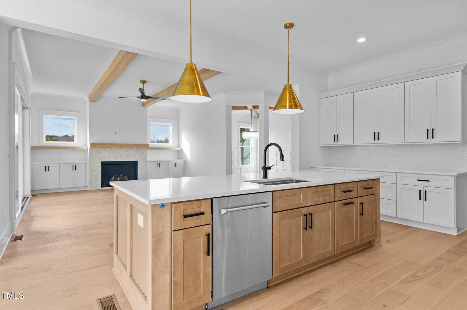Modern kitchen with white walls and wooden flooring featuring a central island with a sink and dishwasher. Two gold pendant lights hang above the island. White cabinets and a wall-mounted oven are on the right, with a fireplace visible in the background.