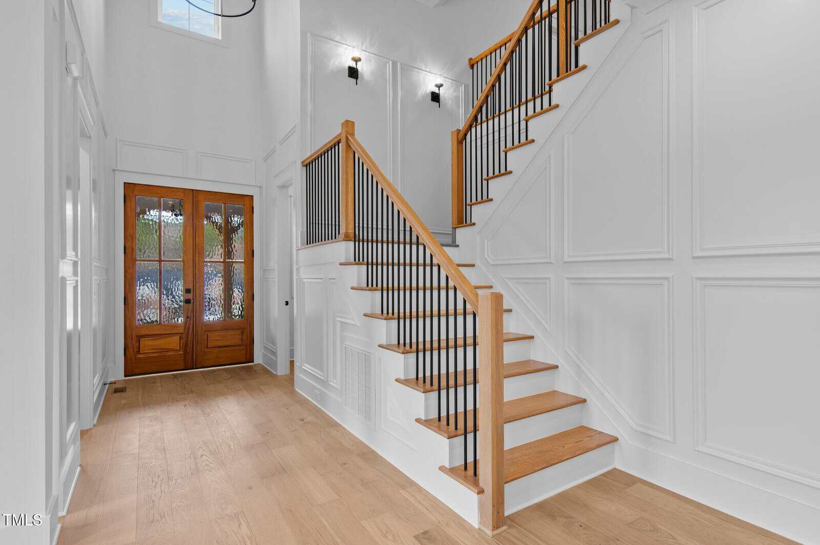 A well-lit foyer featuring a wooden staircase with black metal balusters. The walls are white with wainscoting, and the floor is light wood. Double front doors with glass panels are visible, and two wall lights illuminate the space.