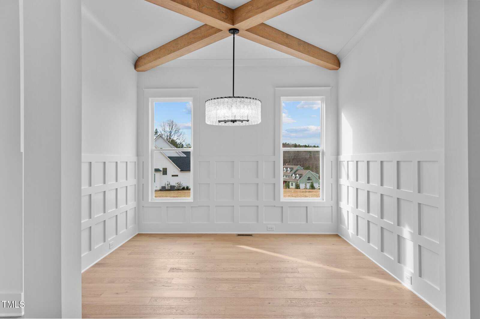 A bright, empty room featuring white walls with a grid pattern, a wooden floor, and a ceiling with exposed wooden beams. Two large windows provide natural light, with a view of houses outside. A modern chandelier hangs from the ceiling.