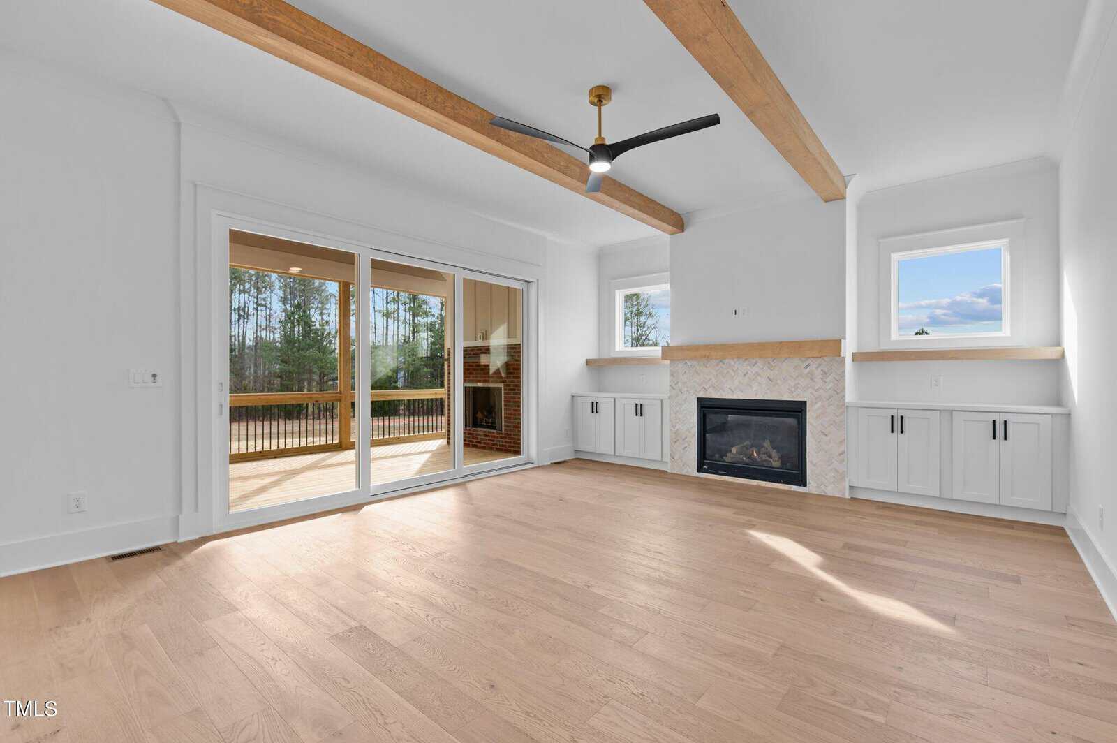 A spacious living room with wooden flooring features a fireplace with a tile surround and built-in cabinets on either side. Large sliding glass doors open to a deck, and a ceiling fan hangs from the ceiling with wooden beams.