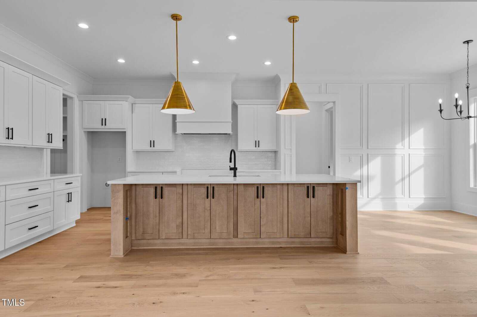 Modern kitchen with white cabinets and a large wooden island featuring a black faucet. Two gold pendant lights hang above the island. The room has wooden flooring, recessed lighting, and a chandelier visible in the adjacent dining area.