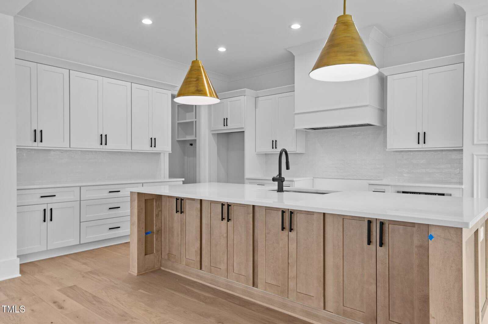 A modern kitchen with white cabinets and a central island featuring light wood panels. Two gold pendant lights hang above the island, and the countertop is white. The faucet is black, and the floor is light wood.