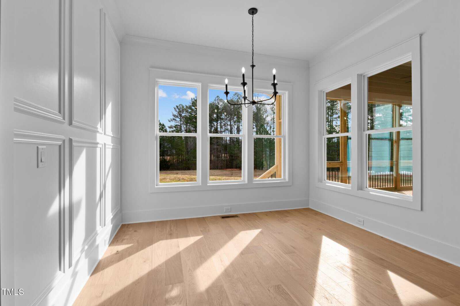 Bright room with light wood flooring and white walls featuring decorative molding. Large windows and a glass door provide views of trees and a patio. A black chandelier hangs from the ceiling, and sunlight streams in, creating shadows on the floor.