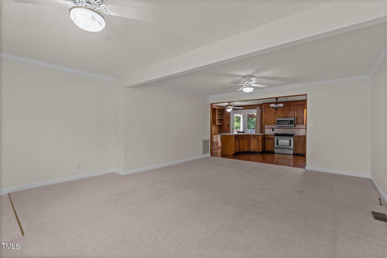 A vacant living room with beige carpet and white walls opens into a kitchen. The kitchen features wooden cabinets, stainless steel appliances, and a ceiling fan. Two ceiling fans with lights are visible in the living room.