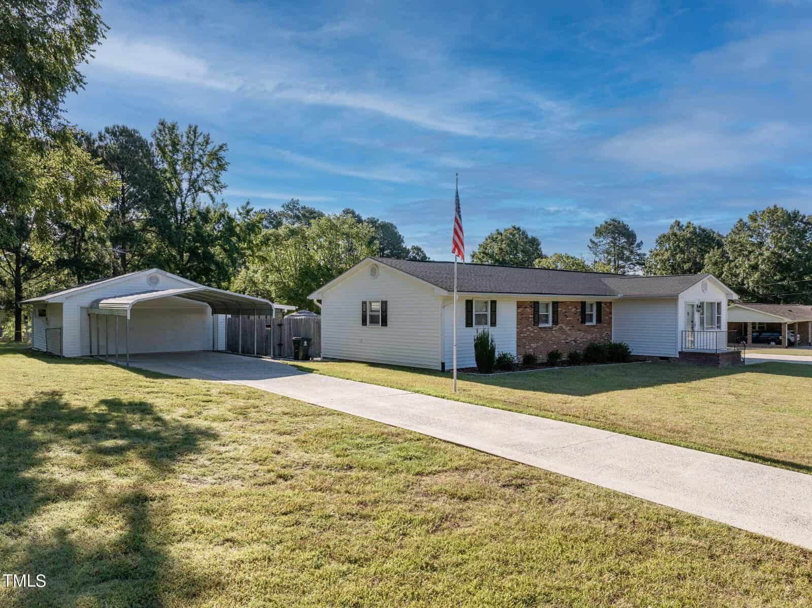 Single-story house with a brick and white exterior, featuring a well-maintained lawn. An American flag is on display in front. Next to the house is a covered carport. Trees and a clear blue sky are visible in the background.