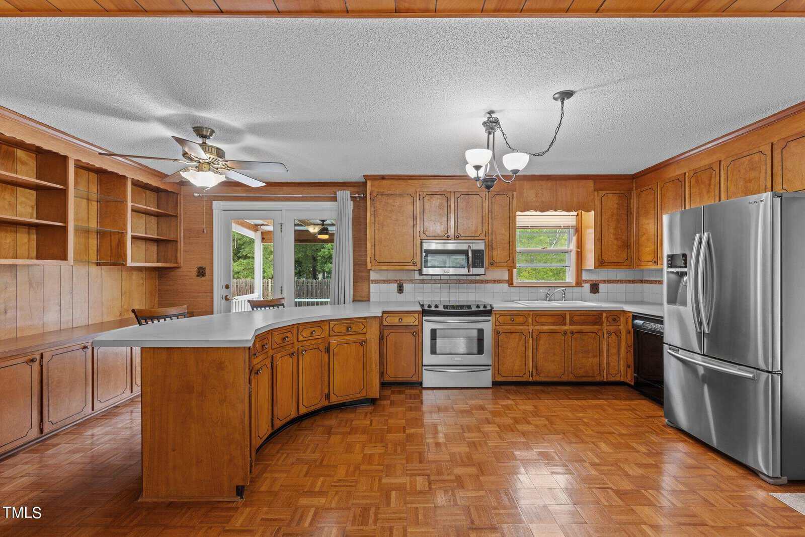 A spacious kitchen with wooden cabinets, stainless steel appliances, and a large island. Parquet flooring is present, along with built-in shelves. A ceiling fan and chandelier provide lighting, and a window offers a view of a green yard.