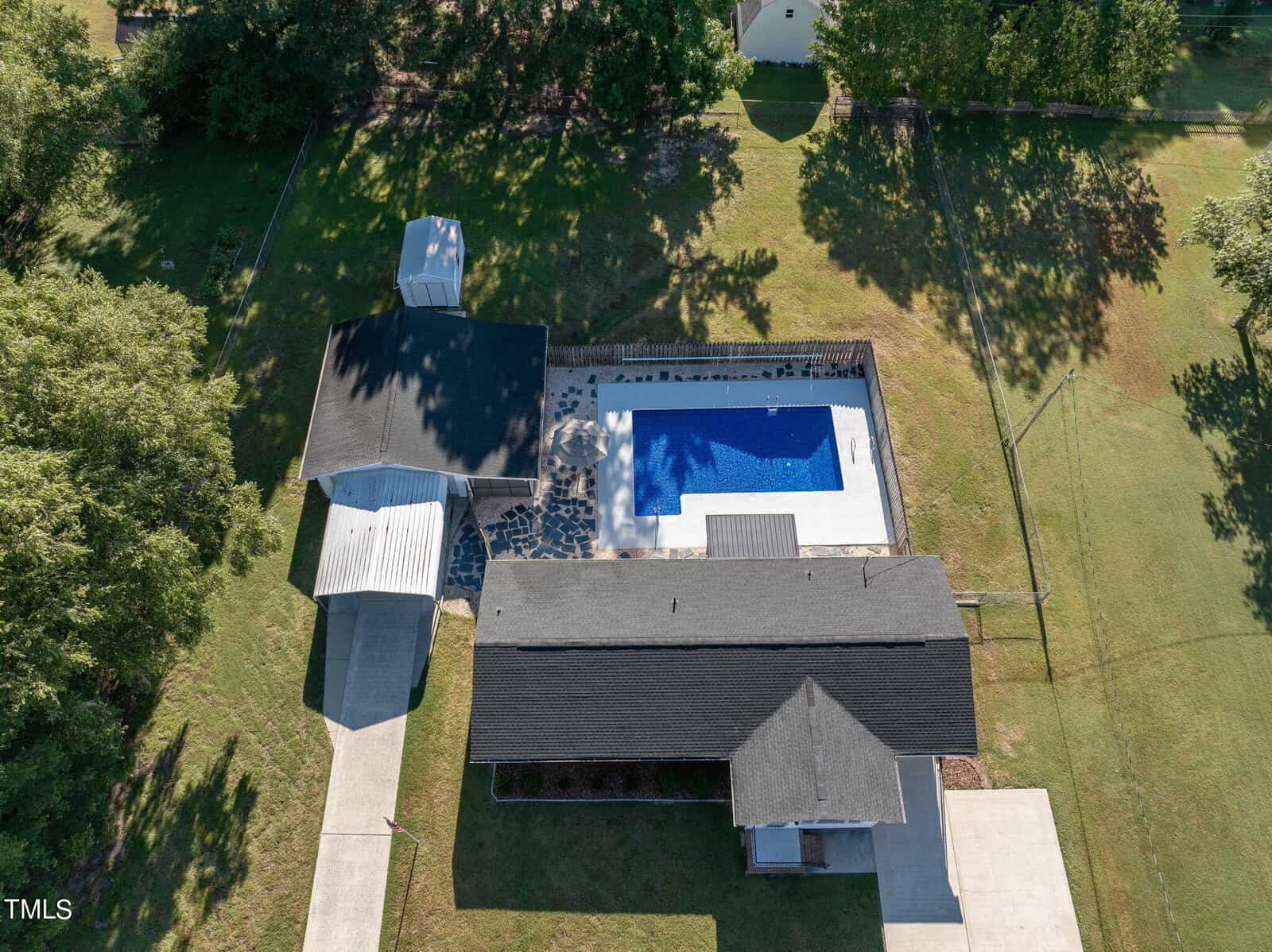 Aerial view of a property with a house, a black-roofed building, and an outdoor swimming pool surrounded by a fence. The grounds have green grass and trees, and a paved driveway leads to the buildings.